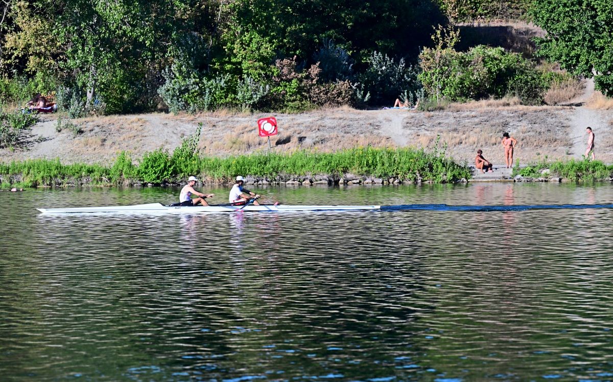 Zwei Personen rudern in einem schmalen weißen Boot über einen Fluss. Im Hintergrund sind eine grasbewachsene Uferböschung, Bäume und Menschen zu sehen, die am Ufer entspannen oder im Wasser sind. Ein Rettungsring-Schild ist sichtbar.