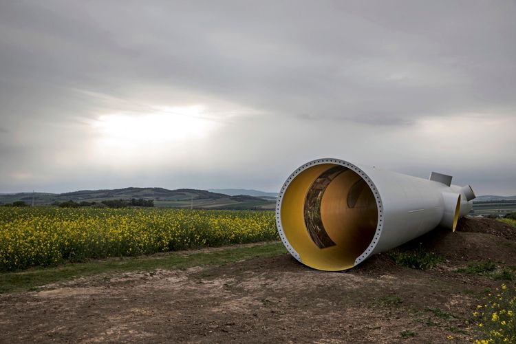 Horizontalturm aus weißem Metall mit gelbem Innenraum, liegend auf einem Feldweg in einer Landschaft mit blühenden gelben Rapsfeldern und sanften Hügeln unter bewölktem Himmel.
