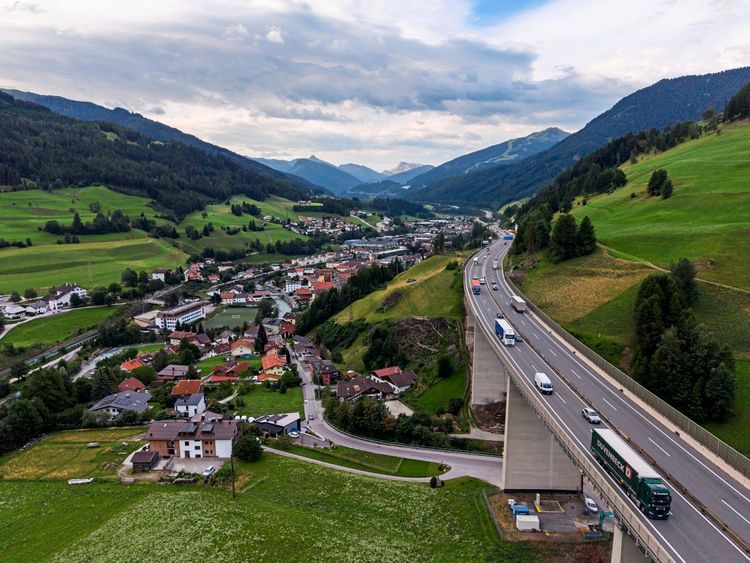 Luftaufnahme einer Autobahnbrücke am Brenner mit Fahrzeugen, umgeben von grünen Hügeln, einer Ortschaft und bewaldeten Bergen im Hintergrund.