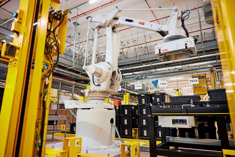 Ein weißer ABB-Industrieroboterarm befindet sich in einem modernen, automatisierten Logistikzentrum mit gelben und schwarzen Strukturelementen. Im Hintergrund sind zahlreiche Regale, Kabel und Beschilderungen zu sehen, die das industrielle Arbeitsumfeld in einem Amazon-Entwicklungszentrum in Vercelli, Italien, zeigen.