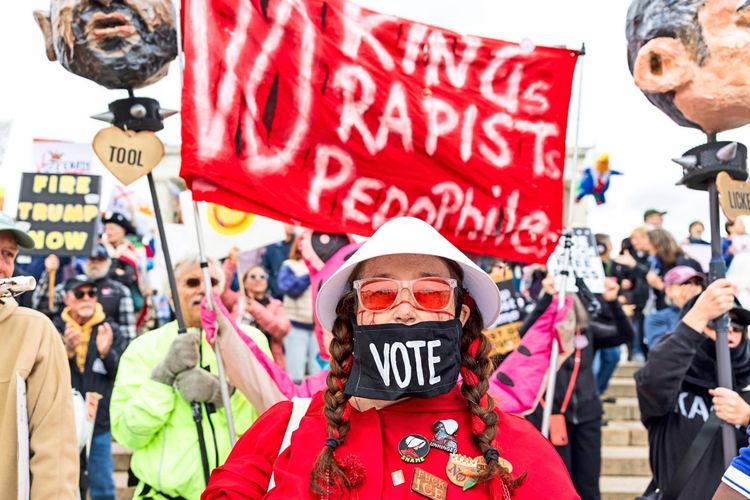 Eine Gruppe von Demonstranten versammelt sich vor dem Lincoln Memorial in Washington, DC, bei einer Kundgebung mit dem Titel „No Kings: Vote Early“. Im Vordergrund steht eine Person mit einer Maske mit der Aufschrift „Vote“, roten Zöpfen und einem weißen Hut. Hinter ihr sind Schilder und Transparente mit politischen Botschaften sowie weitere Teilnehmer sichtbar. Die Kundgebung zielt darauf ab, zur frühen Abstimmung bei den Zwischenwahlen und zur Sicherstellung des Zugangs zu Wahllokalen aufzurufen.