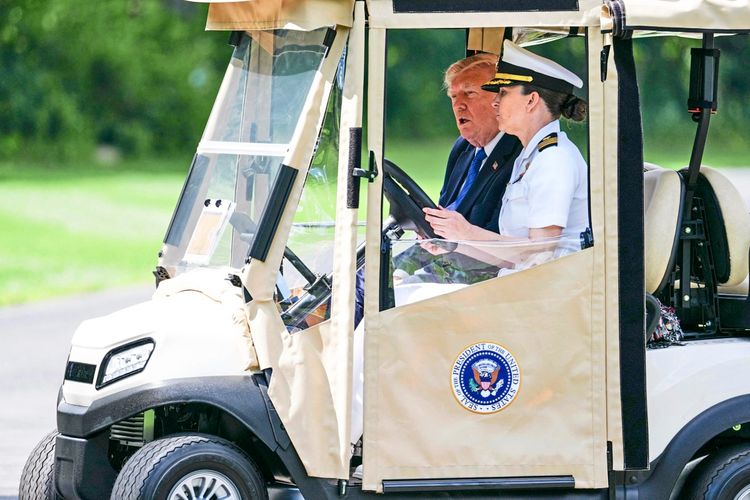 Ein Golfcart mit einem Mann in einem Anzug und einer Uniformierten, die fährt. Das Fahrzeug trägt das Siegel des Präsidenten der Vereinigten Staaten. Im Hintergrund ist eine grüne, bewaldete Umgebung zu sehen.