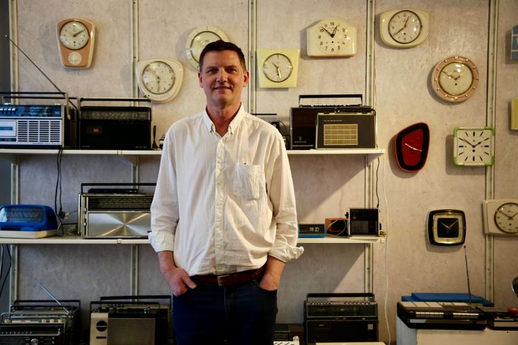 Wolfgang Kremsner vor einem Regal mit alten Kassettenrekordern und einer Wand mit alten Küchenuhren.