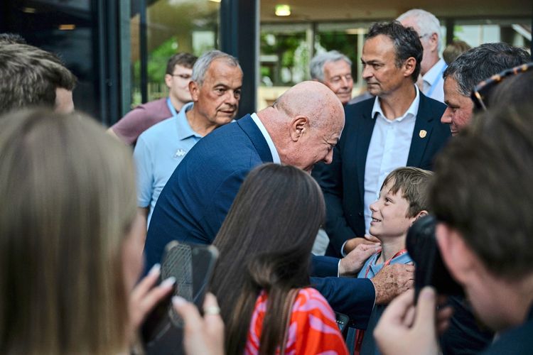 Eine Gruppe von Menschen unter freiem Himmel. Ein älterer Mann mit Glatze spricht lächelnd mit einem Jungen, während andere Personen im Hintergrund stehen und zusehen.