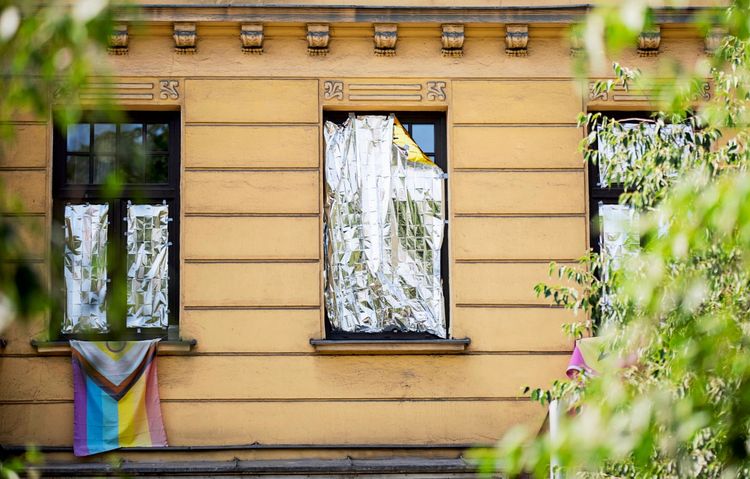 Fenster eines gelben Gebäudes, verhängt mit Alufolie als Schutz vor Sommerhitze. An einem Fenster hängt eine Progress-Pride-Flagge. Im Vordergrund sind teils unscharfe Äste zu sehen.