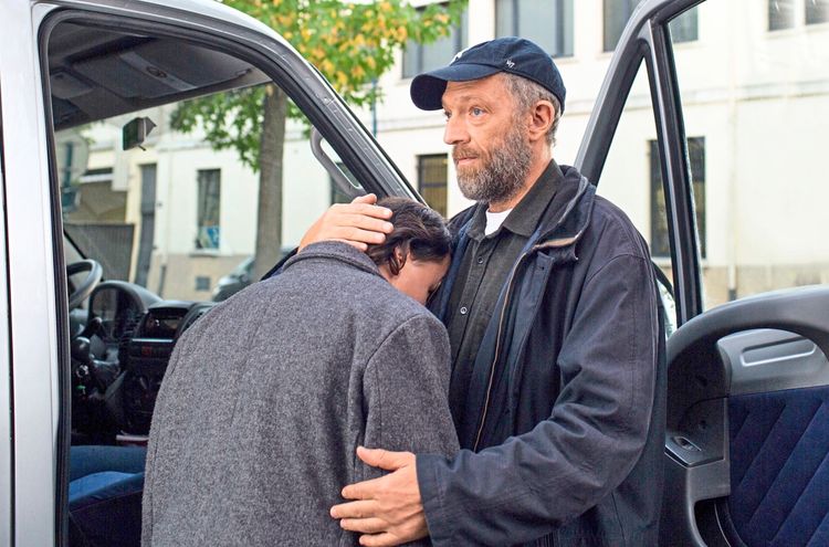 Ein Mann mit Bart und Baseballkappe umarmt eine Person, die ihren Kopf an seine Brust lehnt. Sie stehen neben einem geöffneten Autotür, im Hintergrund sind Gebäude und Bäume sichtbar.