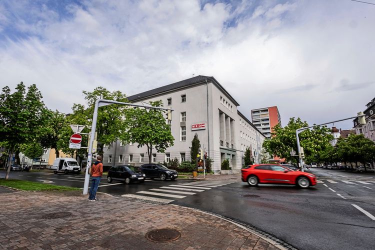 Straßenszene mit einem großen, weißen Gebäude der Wirtschaftskammer (WKO) im Hintergrund. Ein rotes Auto fährt im Vordergrund, während ein Fußgänger eine Ampel überquert. Die Umgebung ist von Bäumen gesäumt, und der Boden wirkt von Regen nass.
