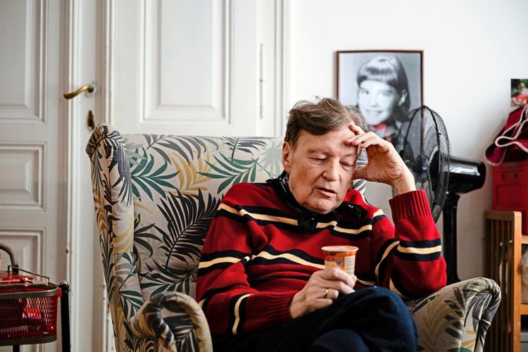 Ein Mann in einem gestreiften roten Pullover sitzt nachdenklich in einem Sessel und hält eine kleine Dose in der Hand. Im Hintergrund ist ein gerahmtes Foto eines Kindes zu sehen.