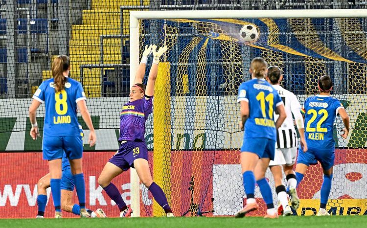 Eine Szene aus einem Fußballspiel: Die Torhüterin im lila Trikot versucht, den Ball abzuwehren, während er ins Tor fliegt. Mehrere Spielerinnen im blauen und weißen Trikot sind im Strafraum zu sehen. Hintergrund: ein Fußballtor mit gelb-blauem Netz und Tribünen.