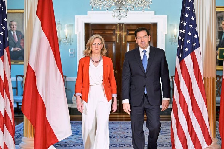 Zwei Personen schreiten in einem formellen Raum zwischen zwei Flaggen (der österreichischen und der US-amerikanischen) auf die Kamera zu. Im Hintergrund sind ein Kronleuchter, Türen und Bilder an den Wänden zu sehen.