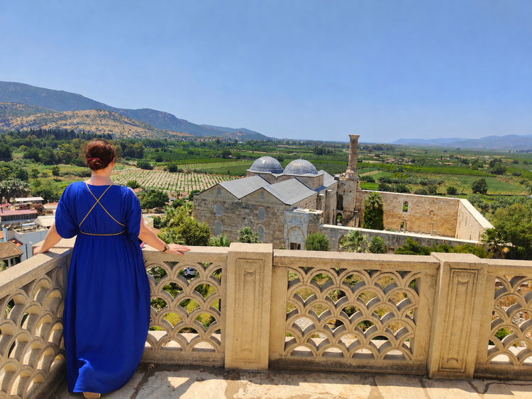 Eine Frau in einem blauen Kleid steht auf einem Balkon mit kunstvoll gestaltetem Geländer. Sie blickt auf eine historische Gebäudeanlage mit Kuppeln und einem hohen Schornstein, umgeben von üppiger grüner Landschaft und Bergen im Hintergrund. Der Himmel ist klar und blau.