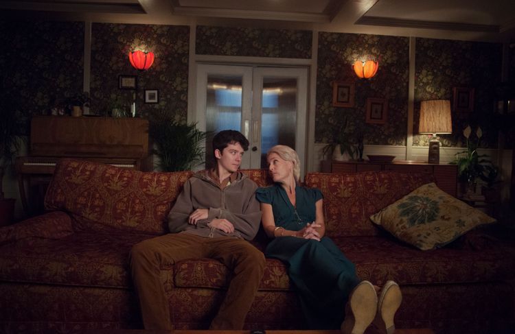 Links: Asa Butterfield als Otis sitzt neben seiner Mutter Jean (Gillian Anderson) auf dem Sofa und blickt sie erwartungsvoll an