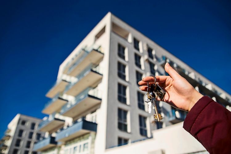 Eine Person hält einen Schlüsselbund mit Wohnungsschlüsseln hoch, im Hintergrund ist ein modernes Wohngebäude mit Balkonen und blauer Himmel zu sehen.