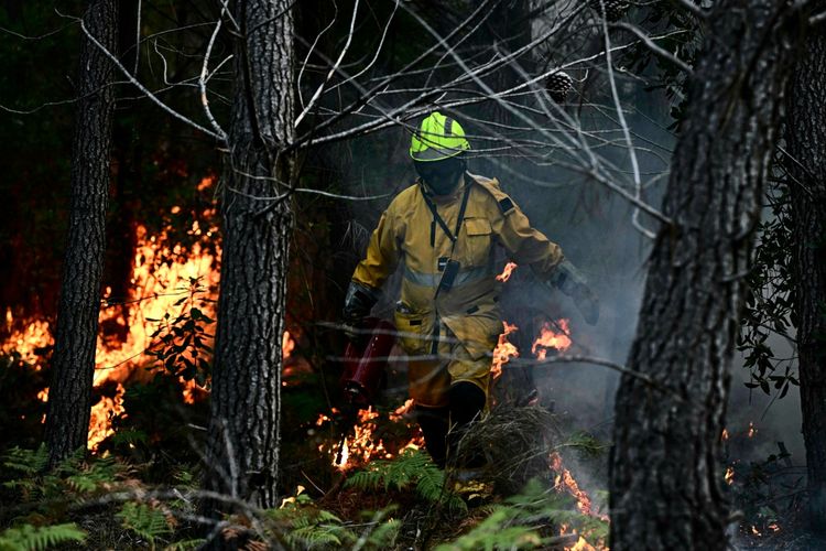 Ein Feuerwehrmann in gelber Schutzkleidung und Helm entzündet mit einer metallenen Drip-Torch-Kanister kontrollierte Feuer in einem Wald in Le Porge, nahe dem Arcachon-Becken, während Brände in der Region Gironde in Südwestfrankreich wüten. Flammen und Rauch sind im Hintergrund sichtbar.