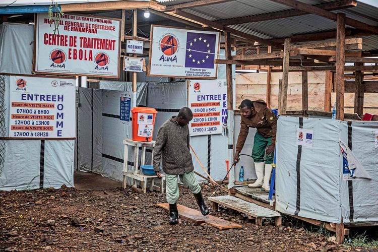 Eine Person desinfiziert die Schuhe eines Mannes am Eingang des Rwampara Ebola-Behandlungszentrums in Bunia, Ituri, im Osten der Demokratischen Republik Kongo. Umgebende Schilder informieren über Zugangszeiten und Sicherheitsmaßnahmen zum Schutz vor der Ebola-Epidemie. Im Hintergrund sind einfache Strukturen und Zelte sichtbar.