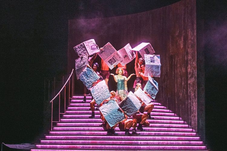 Eine Bühnenperformance mit einer Frau im Zentrum, die ein korsettartiges Kostüm trägt, und von mehreren Tänzern umgeben ist. Sie halten große, gefaltete Zeitungsmotive und stehen auf einer beleuchteten Treppe mit dramatischer Beleuchtung.