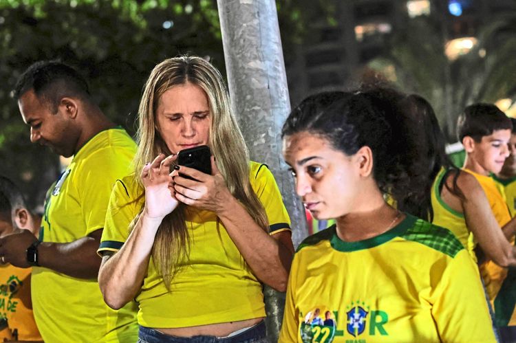 Eine Gruppe von Menschen in gelben Trikots, die die brasilianische Nationalmannschaft repräsentieren. Eine Frau schaut konzentriert auf ihr Smartphone, möglicherweise um Wahlergebnisse zu überprüfen. Im Hintergrund sind weitere Personen zu sehen, die ebenso in gelben Trikots gekleidet sind. Die Szene spielt sich nachts in einer Außenumgebung ab.