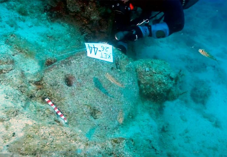 Ein Taucher untersucht unter Wasser eine Ausgrabungsstelle mit Steinankern. Neben einem Steinanker sind zwei Löcher sichtbar, die für Taue verwendet wurden. Eine beschriftete Tafel mit „KET 26-44“ markiert die Fundstelle. Im Hintergrund schwimmen zwei Fische, und am Boden liegt eine rot-weiße Maßstabsstange.