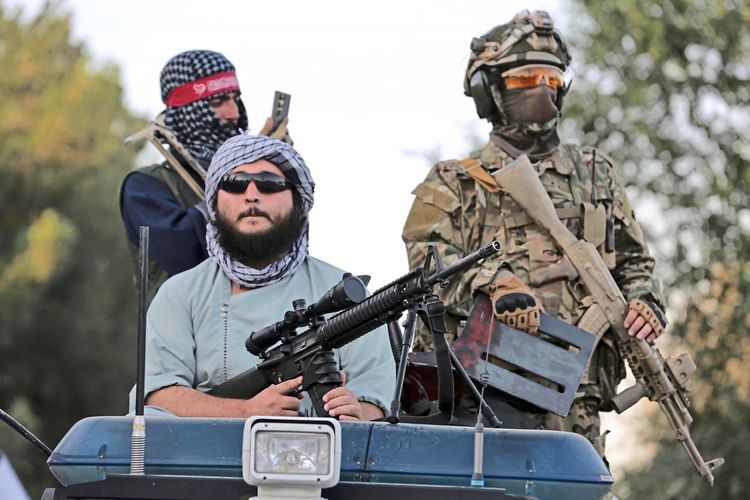 Drei bewaffnete Männer, zwei in militärischer Ausrüstung und einer mit traditioneller Kleidung und Schal, sichern eine Veranstaltung in Kabul, Afghanistan. Die Szene zeigt eine Feier zum fünften Jahrestag der Machtübernahme durch die Taliban am 15. August 2026. Im Hintergrund sind Bäume zu sehen.