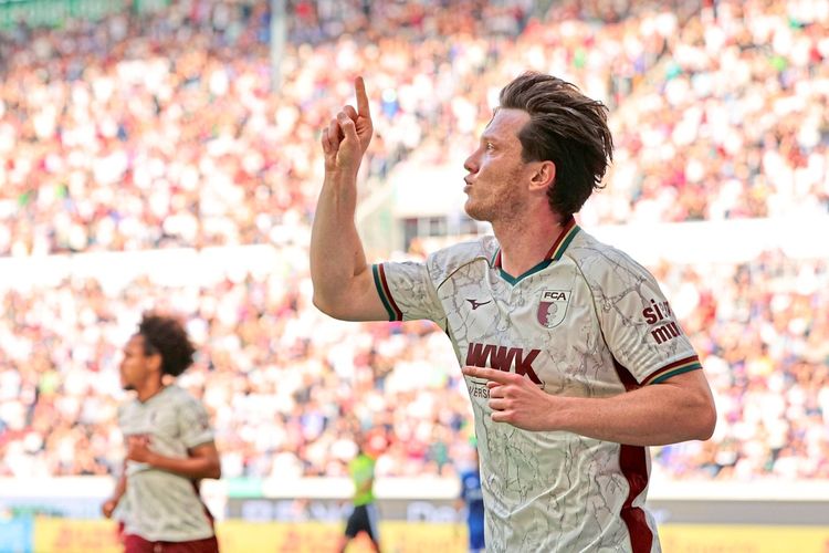 Ein Fußballspieler des FC Augsburg in weißem Trikot jubelt mit erhobenem Zeigefinger nach einem Tor während eines Bundesliga-Spiels gegen den FC Schalke 04 in Augsburg, Deutschland. Im Hintergrund sind unscharfe Zuschauer und ein Mitspieler zu sehen.