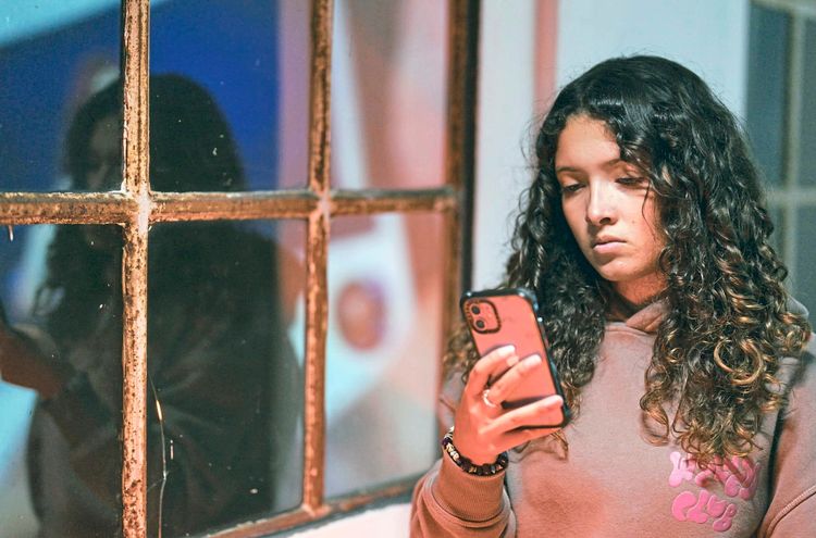 Eine Frau mit lockigem Haar schaut ernst auf ihr Smartphone, während sie in einem Raum steht. Im Hintergrund ist ihr Spiegelbild in einem alten, rostigen Fenster erkennbar.