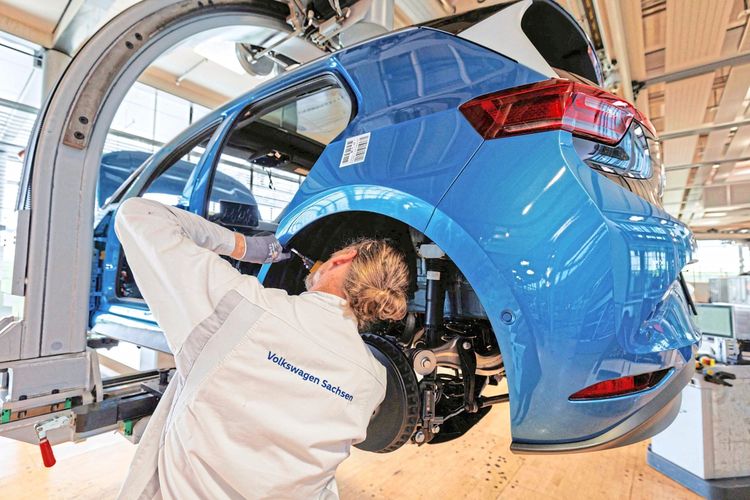 Ein Mitarbeiter in weißer Arbeitskleidung mit der Aufschrift „Volkswagen Sachsen“ arbeitet an der Montage eines blauen Volkswagen ID.3 Elektroautos in der „Gläsernen Manufaktur“ in Dresden. Das Fahrzeug befindet sich auf einer Hebevorrichtung.