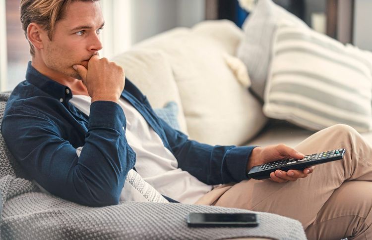 Ein Mann sitzt mit einer Fernbedienung in der Hand auf der Couch und schaut kritisch Richtung Fernseher