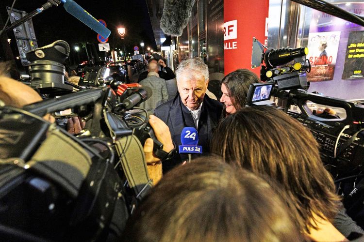 Eine Person wird von mehreren Mikrofonen und Kameras bei einer Filmpremiere im Gartenbaukino in Wien umringt. Im Hintergrund sind ein rotes Poster und eine Filmankündigung sichtbar.