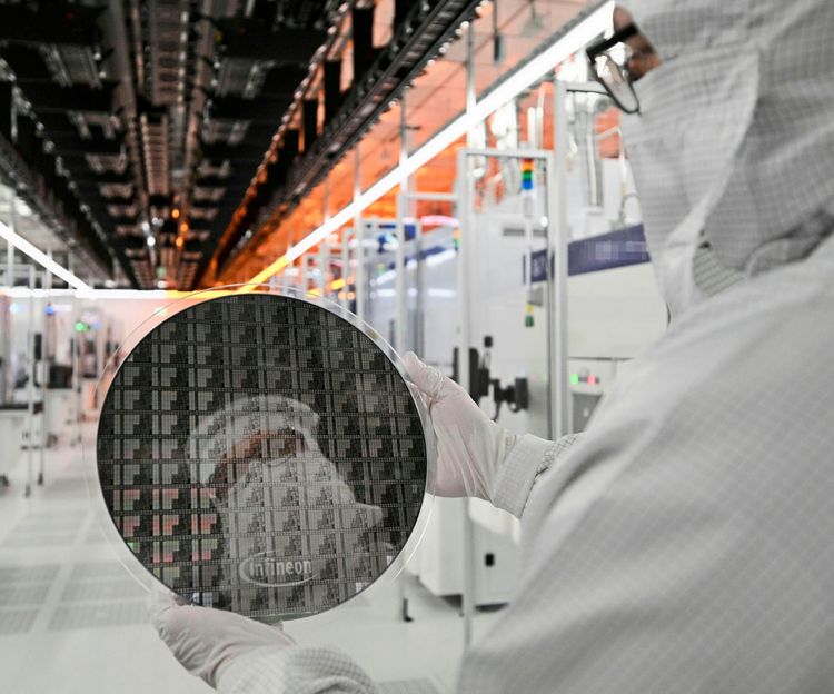 Ein Mitarbeiter in Schutzanzug und Handschuhen spiegelt sich in einem 300-mm-Galliumnitrid-Wafer, den er in einem vollautomatisierten Reinraum einer Chipfabrik hält. Im Hintergrund sind technische Einrichtungen und Beleuchtungen sichtbar.