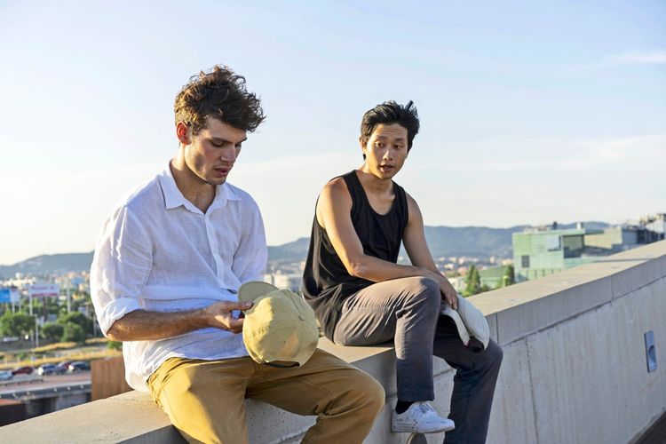 Zwei junge Männer sitzen auf einer Betonmauer und unterhalten sich. Einer trägt ein weißes Hemd und hält eine beige Mütze, der andere trägt ein schwarzes Tanktop und eine graue Hose mit einer Mütze in der Hand. Im Hintergrund sind eine Landschaft mit Hügeln und moderne Gebäude zu sehen.