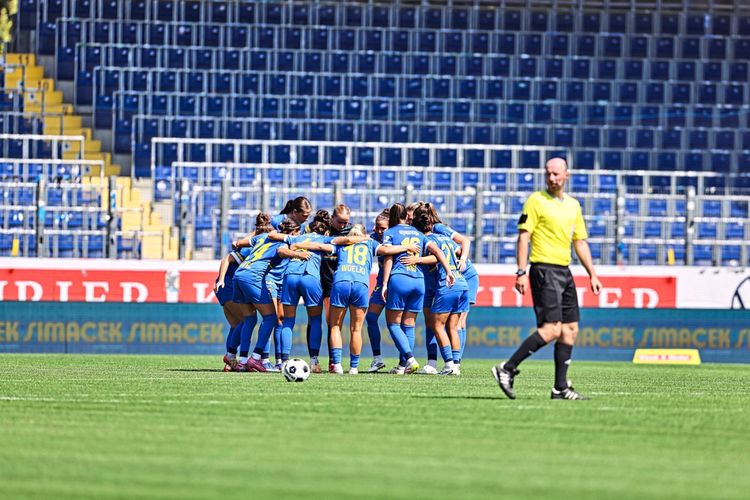Eine Frauenfußballmannschaft von SKN St. Pölten bildet vor einem Spiel in der Bundesliga gegen Altach einen Mannschaftskreis auf dem Spielfeld. Im Vordergrund ist ein Schiedsrichter zu sehen. Im Hintergrund leere Stadiontribünen.