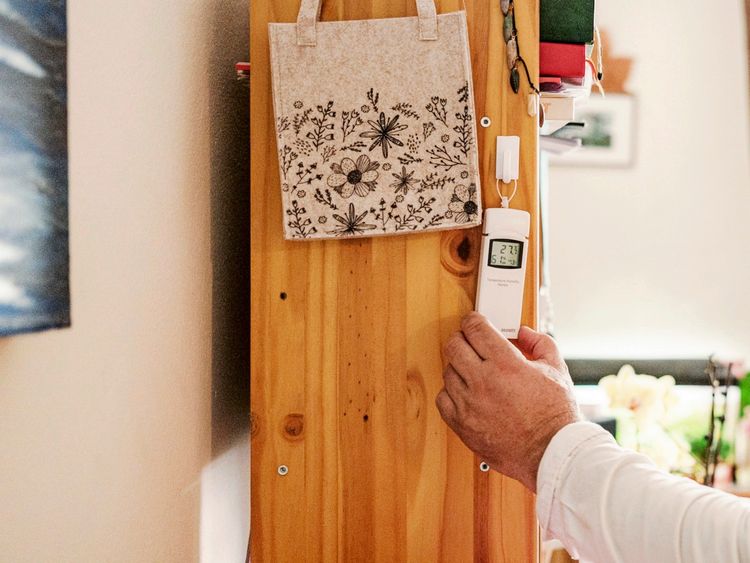 Ein Holzregal in einem Raum, an dem eine kleine Stofftasche mit Blumenmotiv hängt. Darunter misst eine Hand mit einem digitalen Thermometer die Raumtemperatur, die auf dem Gerät mit 27,3 °C und einer Luftfeuchtigkeit von 56 % angezeigt wird. Im Hintergrund sind unscharf Blumen und weitere Wohnaccessoires zu erkennen.