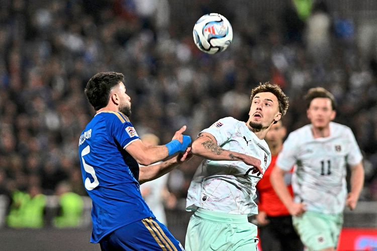 Zwei Fußballspieler, einer in blauem und einer in weißem Trikot, kämpfen während eines Spiels in einem Stadion um den Ball, der in der Luft schwebt. Zuschauer sind im Hintergrund sichtbar.