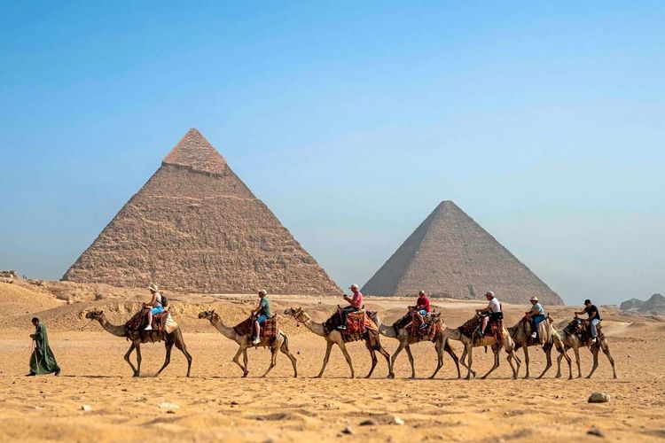 Sieben Kamele, auf denen Touristinnen und Touristen sitzen, werden vor der Chephren- und der Cheops-Pyramide vorbeigeführt.