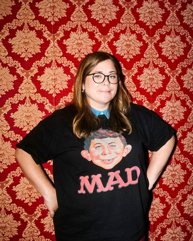 Eine Person mit langen braunen Haaren und Brille steht vor einer roten Tapete mit goldenen Ornamenten. Sie trägt ein schwarzes T-Shirt mit dem Aufdruck „MAD“ und einem Comic-Gesicht.