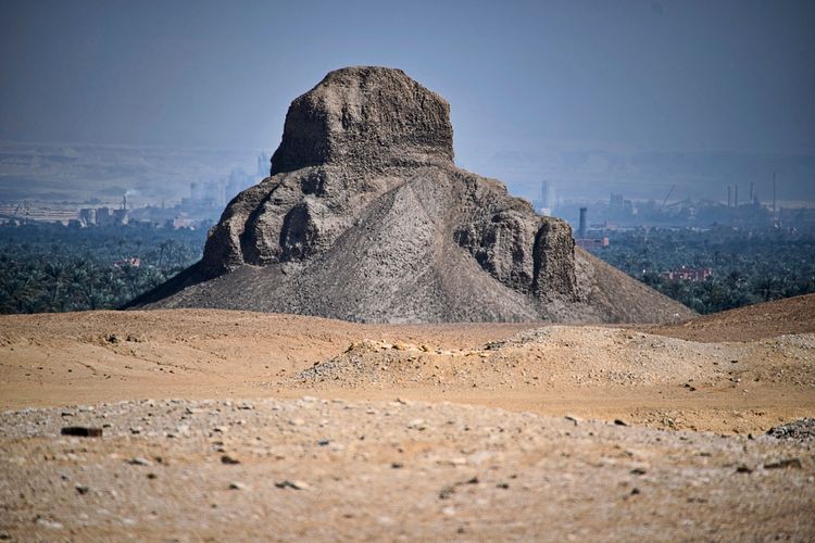 Eine pyramidale Ruine im ägyptischen Wüstengebiet, umgeben von Sand und Vegetation im Hintergrund, mit verschwommenen Gebäuden und Palmen in der Ferne.