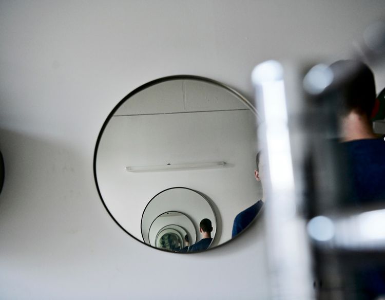 Ein runder Spiegel reflektiert mehrfach ein verschwommenes Bild, das die Rückansicht einer Person zeigt. Die Szene wirkt ruhig und nachdenklich. Im Hintergrund ist eine helle Wand mit einer Lichtleiste zu sehen.