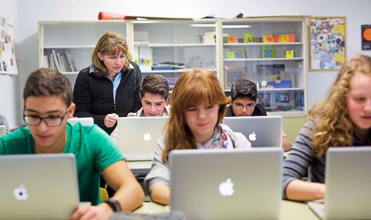 Schüler und Schülerinnen schauen in Laptop, Lehrerin steht zwischen den Reihen.