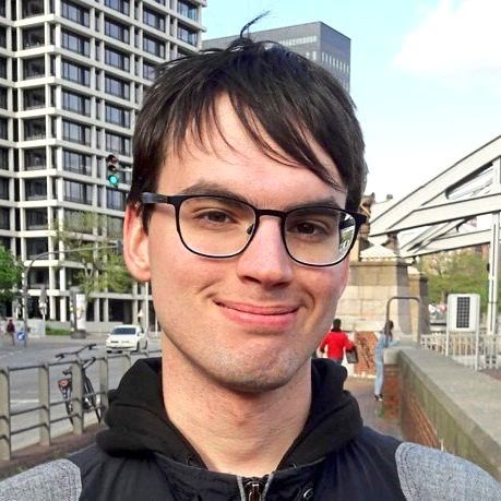 Ein junger Mann mit dunkelbraunem Haar, Brille und einem schüchternen Lächeln steht im Freien vor einem modernen Gebäude und einem Gehweg mit Passanten. Im Hintergrund sind Fahrräder, Autos und städtische Strukturen sichtbar.