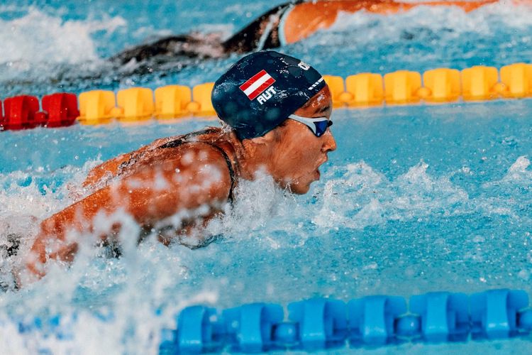 Eine Schwimmerin mit Badekappe und Schwimmbrille im Wettkampf, umgeben von spritzendem Wasser in einem Schwimmbecken. Im Hintergrund sind gelbe und rote Begrenzungslinien im Wasser zu sehen.
