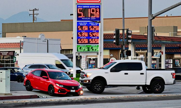 Eine rote Limousine und ein weißer Pickup fahren an einer Tankstelle in La Puente, Kalifornien, vorbei. Im Hintergrund befindet sich eine Preistafel mit angegebenen Kraftstoffpreisen. Ein Einkaufszentrum ist ebenfalls im Hintergrund zu sehen.