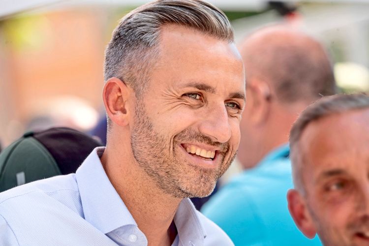 Eine lächelnde Person in einem hellblauen Hemd bei einer Veranstaltung im Freien, mit unscharfem Hintergrund, wo weitere Menschen zu sehen sind.