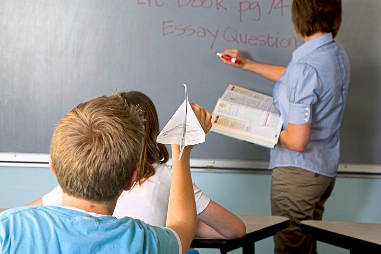 Eine an der Tafel stehende Lehrerin, auf deren Rücken ein Schüler mit einem Papierflieger zielt.