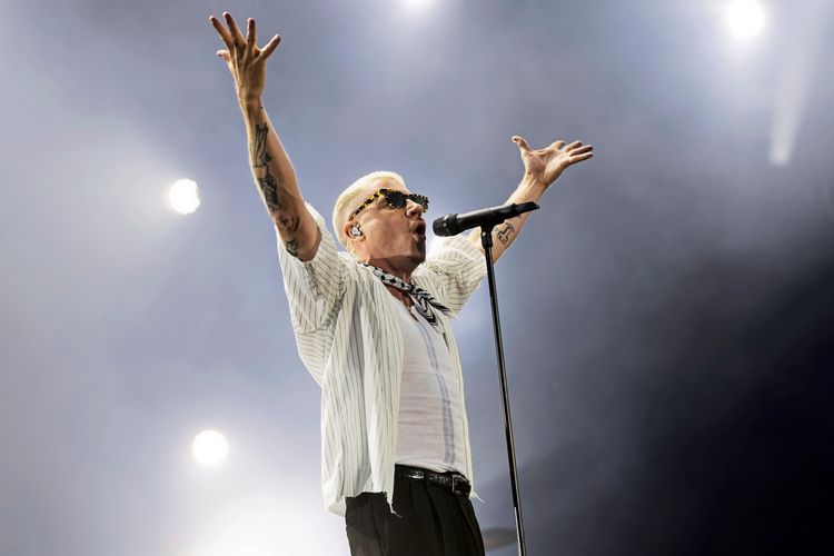 Ein Sänger mit kurzen blonden Haaren und Sonnenbrille performt mit ausgebreiteten Armen auf einer Bühne, beleuchtet von Scheinwerfern, während eines Festivals.