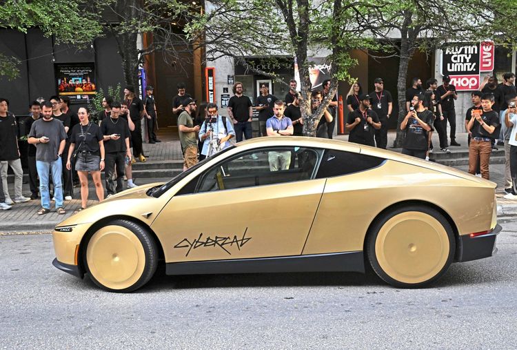 Ein goldfarbenes Tesla-Cybercab-Elektroauto mit der Aufschrift „Cybercab“ an der Seite steht auf einer Straße in Austin, Texas. Im Hintergrund stehen Zuschauer, die das Auto mit ihren Smartphones fotografieren. Die Szene spielt vor dem Veranstaltungsort „Austin City Limits Live“.