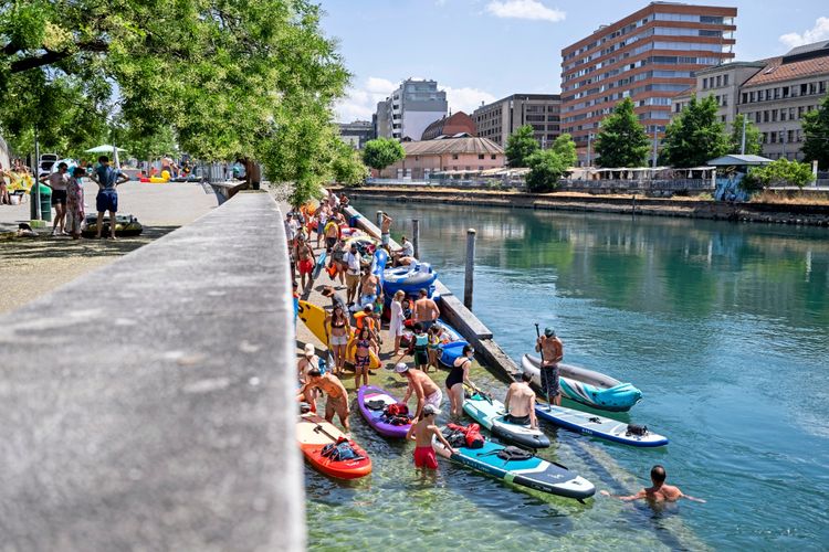 Menschen schwimmen und nutzen Schlauchboote sowie Stand-up-Paddle-Boards im Rhône-Fluss in Genf, Schweiz. Am Ufer sind weitere Personen und Boote zu sehen. Im Hintergrund befinden sich Gebäude und Bäume unter strahlendem Sommerhimmel.