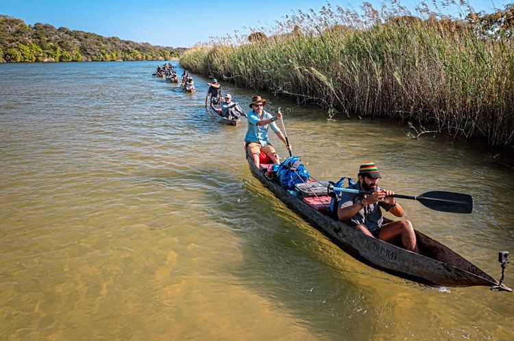 Eine Gruppe von Menschen auf einem Fluss, jeweils zwei Personen sitzen in einem länglichen Ruderboot aus Holz