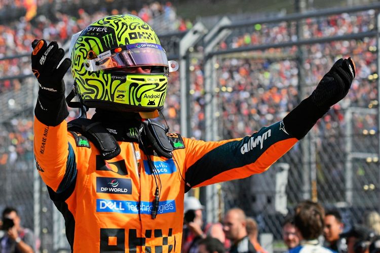 Ein Rennfahrer in einem orangefarbenen McLaren-Rennanzug und einem Helm mit gelbem Design feiert gestikulierend nach einem Rennen auf einer Rennstrecke, umgeben von Zuschauern und Teammitgliedern.