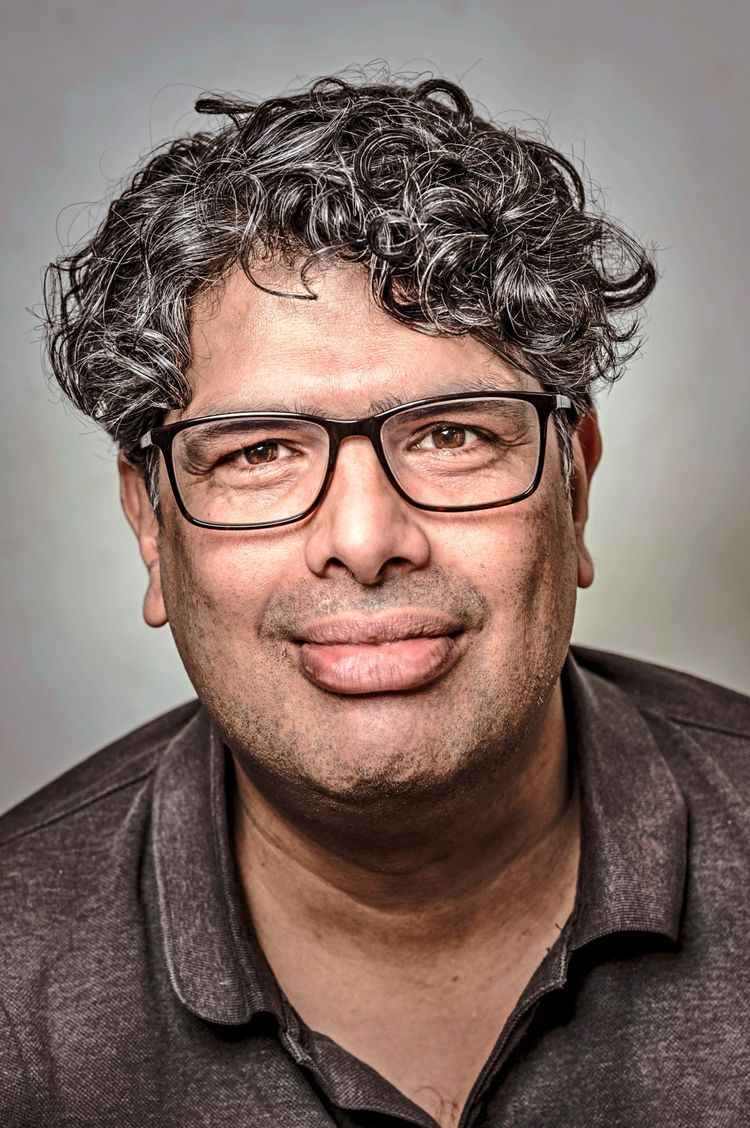 Der Autor Manik Sarkar mit welligem dunklem Haar, Brille und einem dunklen Poloshirt blickt direkt in die Kamera. Der Hintergrund ist neutral und weich, wodurch der Fokus auf dem Gesicht liegt.