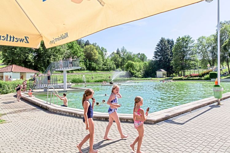 Ein Außenbereich eines Naturfreibades an einem sonnigen Tag. Kinder in Badeanzügen laufen über das gepflasterte Gelände, während andere Personen im Wasser schwimmen oder auf einem Sprungturm stehen. Im Hintergrund sind Bäume, ein Gebäude und ein Wasserbrunnen im Schwimmbecken zu sehen. Im Vordergrund ragt ein Sonnenschirm ins Bild.